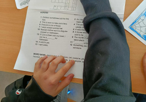 Uczniowie siedzą przy ławkach i wspólnie rozwiązują krzyżówkę z hasłami dotyczącymi Halloween.