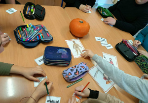 Grupa uczniów siedzi przy stoliku i bierze udział w grach językowych związanych z Halloween, układając karty i wykonując zadania w języku angielskim.